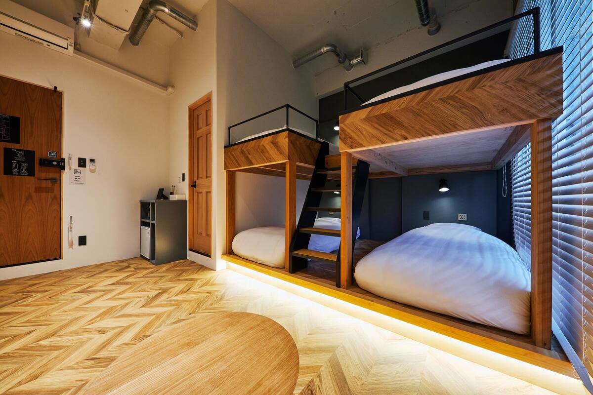 Une chambre avec des lits superposés, un parquet à motifs en chevrons, une petite table et un coin cuisine.