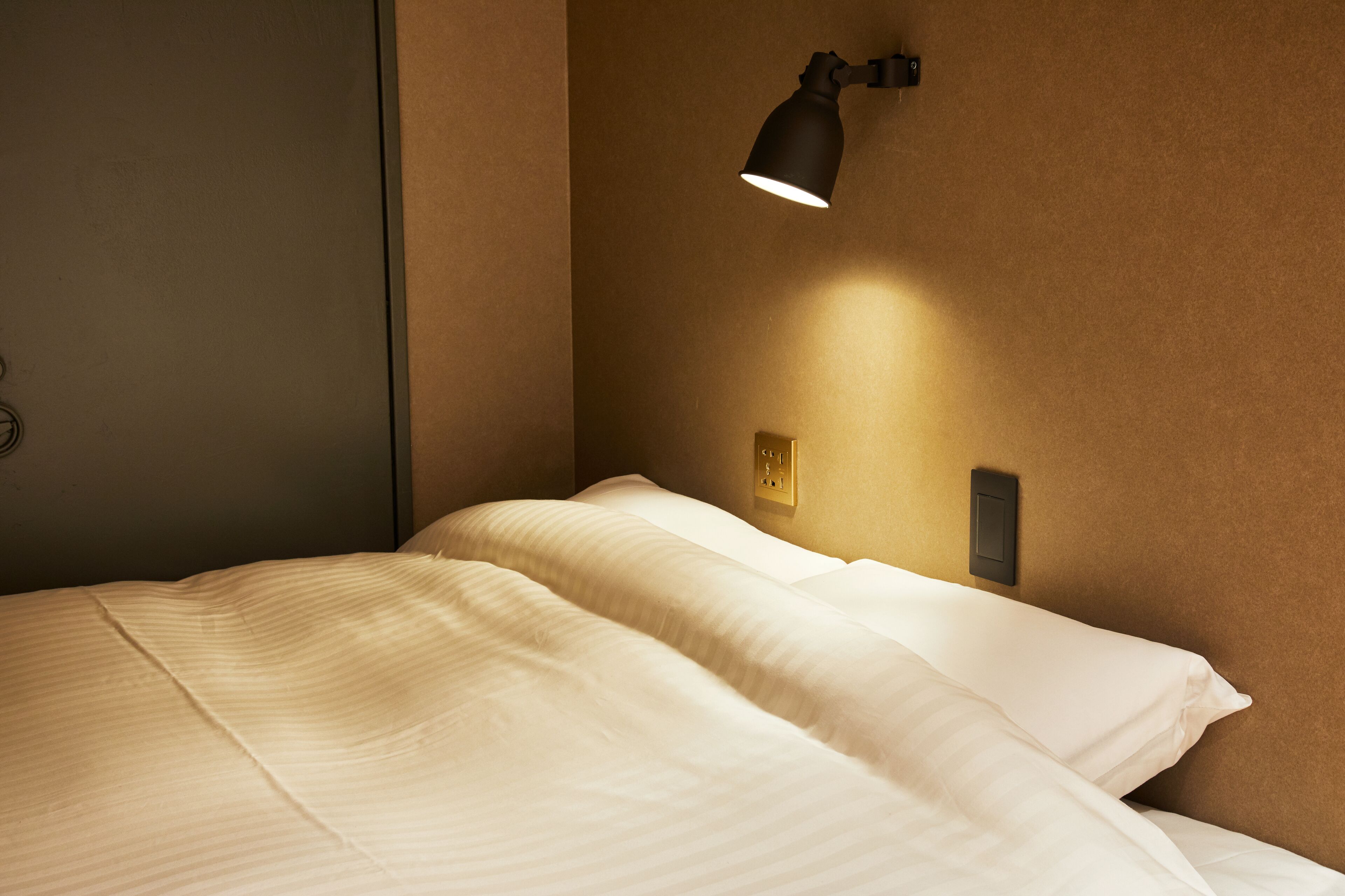 Ein ordentlich bezogenes Bett mit weißen Laken, eine Wandlampe und eine Steckdose an der Wand.