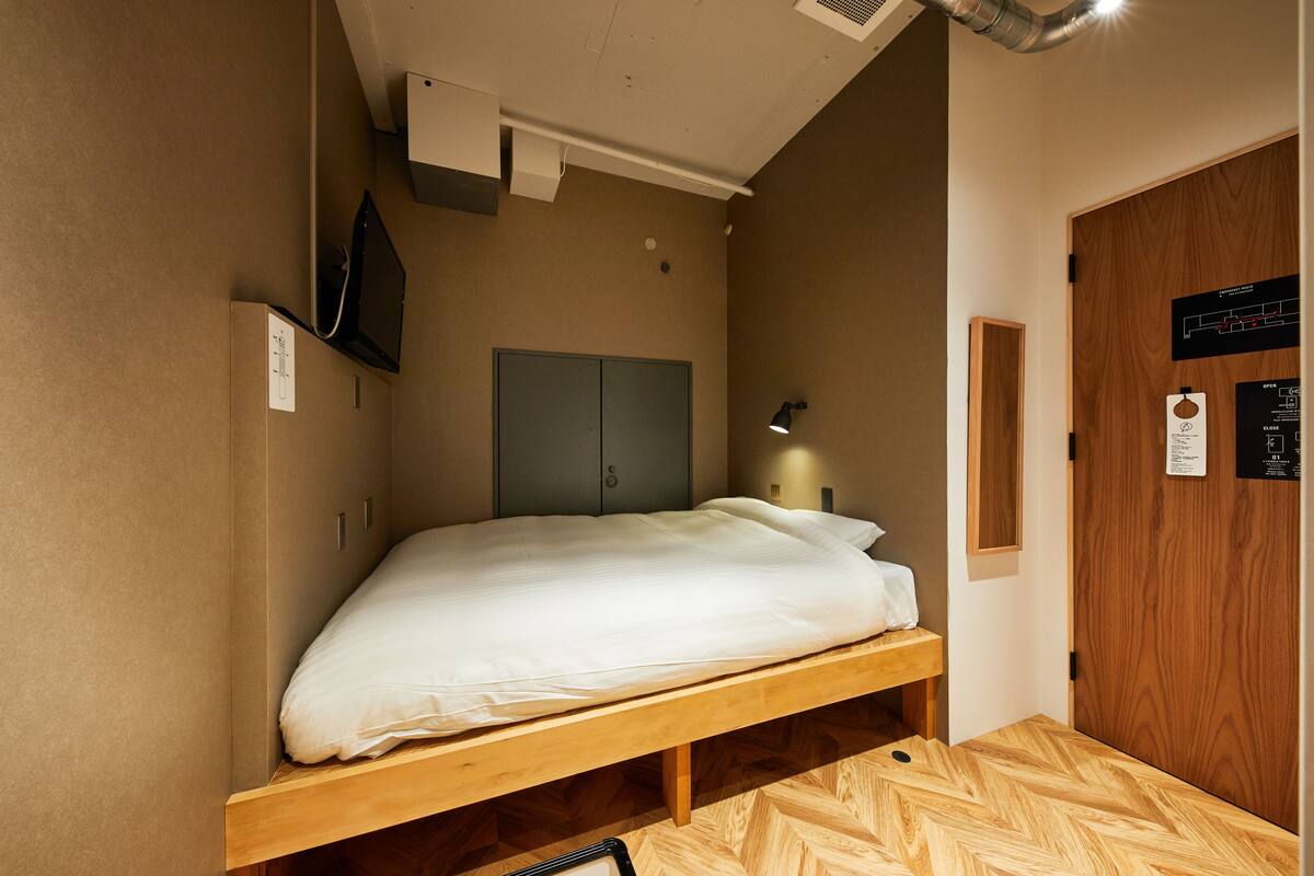 Un lit simple dans une chambre avec un plancher en bois, un téléviseur fixé au mur, une bouche d’aération au plafond et une porte avec un panneau en bois.