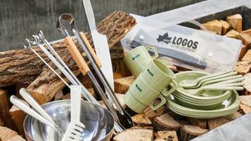 A collection of cutlery, a metal bowl, and a green mug set on a pile of wood logs.