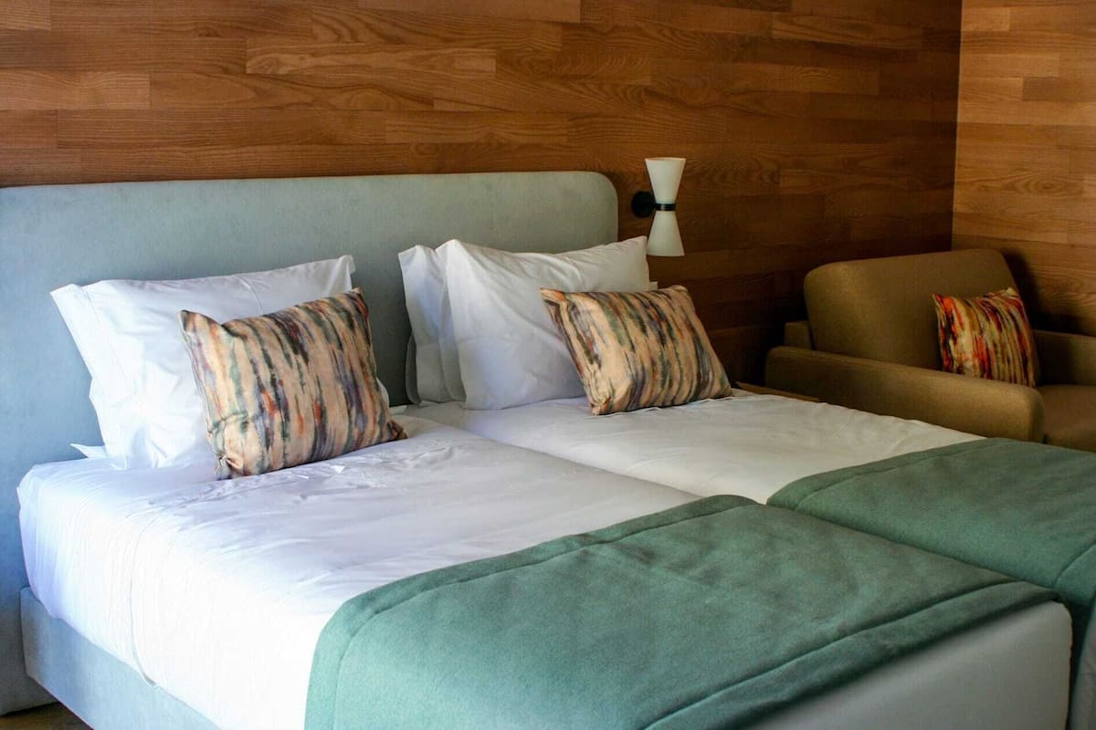 Una cama doble con sábanas blancas y almohadas a rayas, una manta verde y un sillón marrón con un cojín de colores.