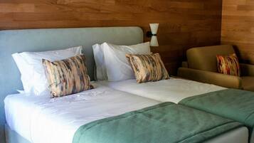 A double bed with white linens and striped pillows, a green throw blanket, and a brown armchair with a colorful pillow.