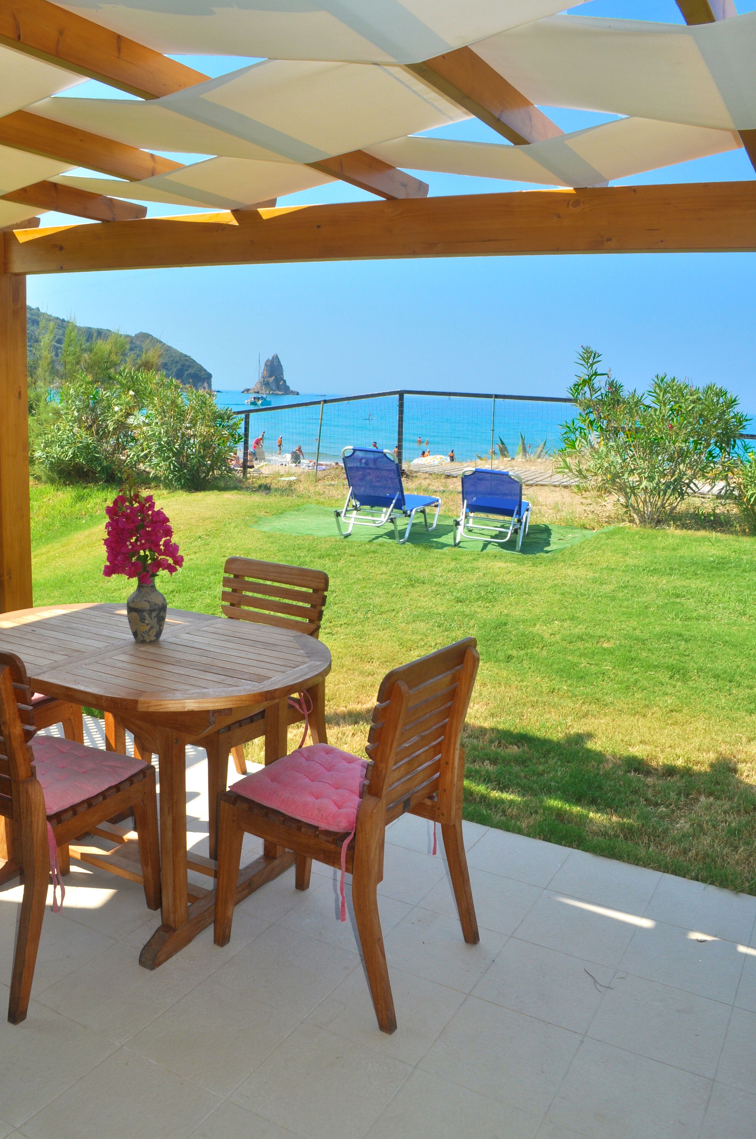 A covered patio with a wooden table and chairs, a vase of flowers, and a view of the beach and ocean.