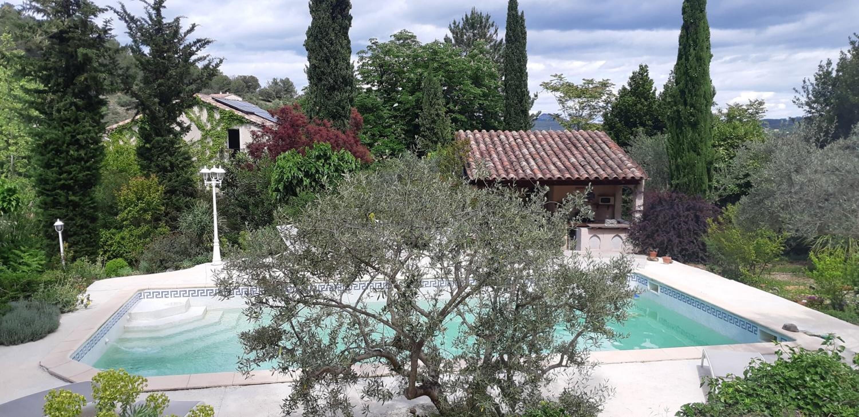 Ein Pool, umgeben von Olivenbäumen und hoch aufragenden Zypressen, mit einem Gebäude mit Ziegeldach im Hintergrund.