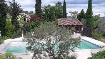 Ein Pool, umgeben von Olivenbäumen und hoch aufragenden Zypressen, mit einem Gebäude mit Ziegeldach im Hintergrund.