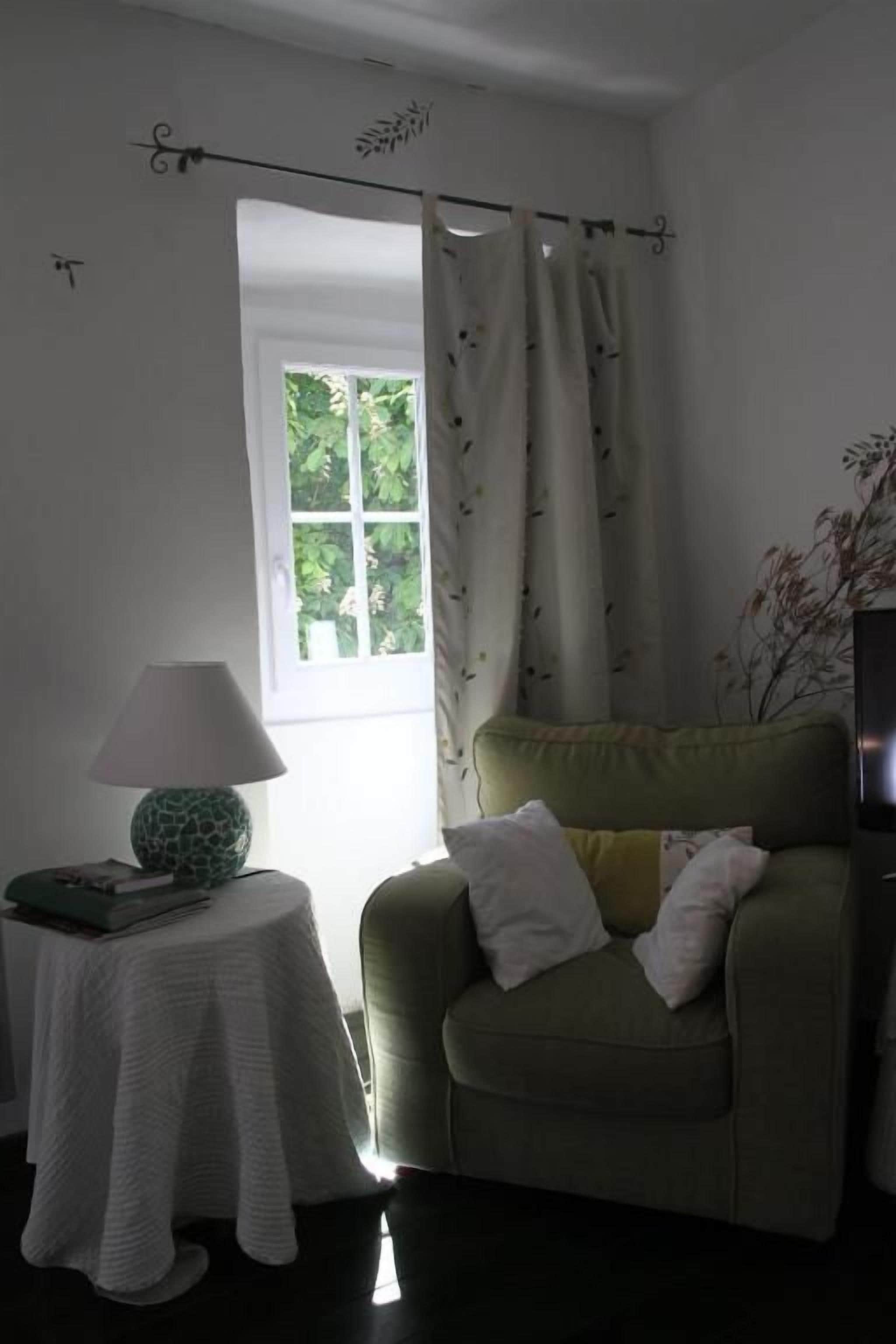 A room with a window, a green armchair with cushions, a small round table with a lamp, and a white tablecloth.