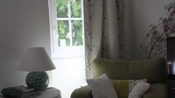 A room with a window, a green armchair with cushions, a small round table with a lamp, and a white tablecloth.