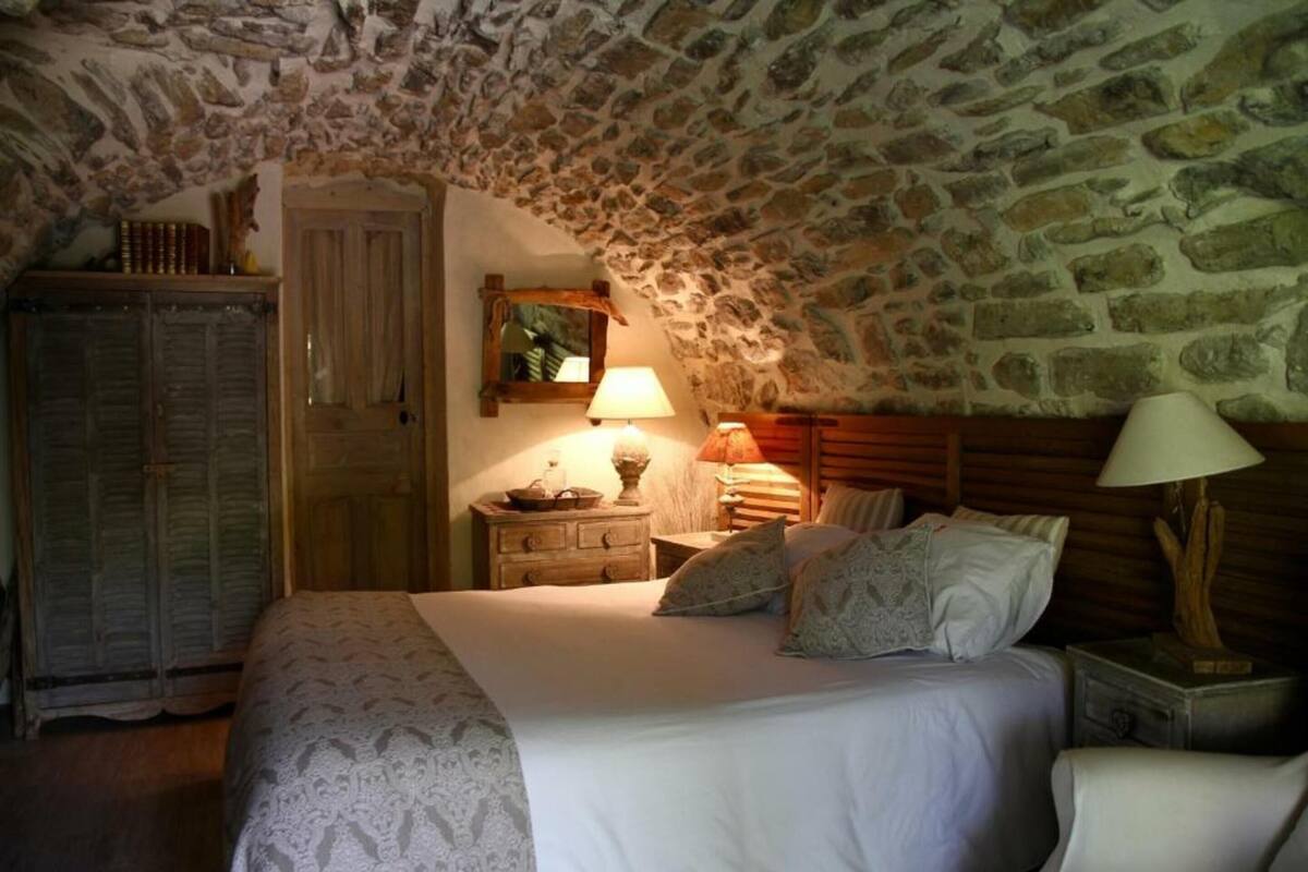 A bedroom with a stone ceiling, a wooden wardrobe, a bedside table with a lamp, and a bed with pillows.