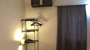 Habitación de hotel con cama, ventilador de techo, unidad de aire acondicionado, lámpara, estante con ropa colgada y ventana con cortinas.