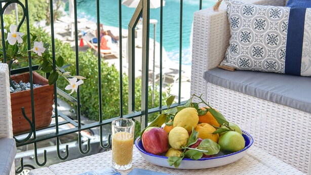 A balcony with wicker furniture, a bowl of fruit, a glass of juice, and a magazine. - Les Suites des Tout le Monde (Sorrento)