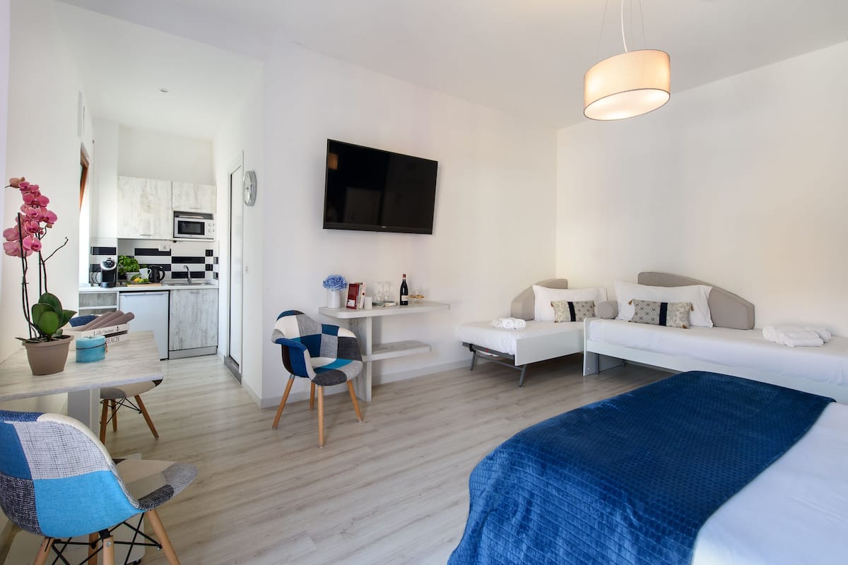 Een moderne hotelkamer met een bed, een bureau, een stoel, een televisie en een kitchenette op de achtergrond.