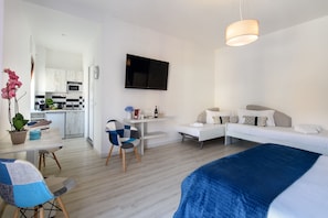 A modern hotel room with a bed, a desk, a chair, a TV, and a kitchenette in the background. - Les Suites des Tout le Monde (Sorrento)