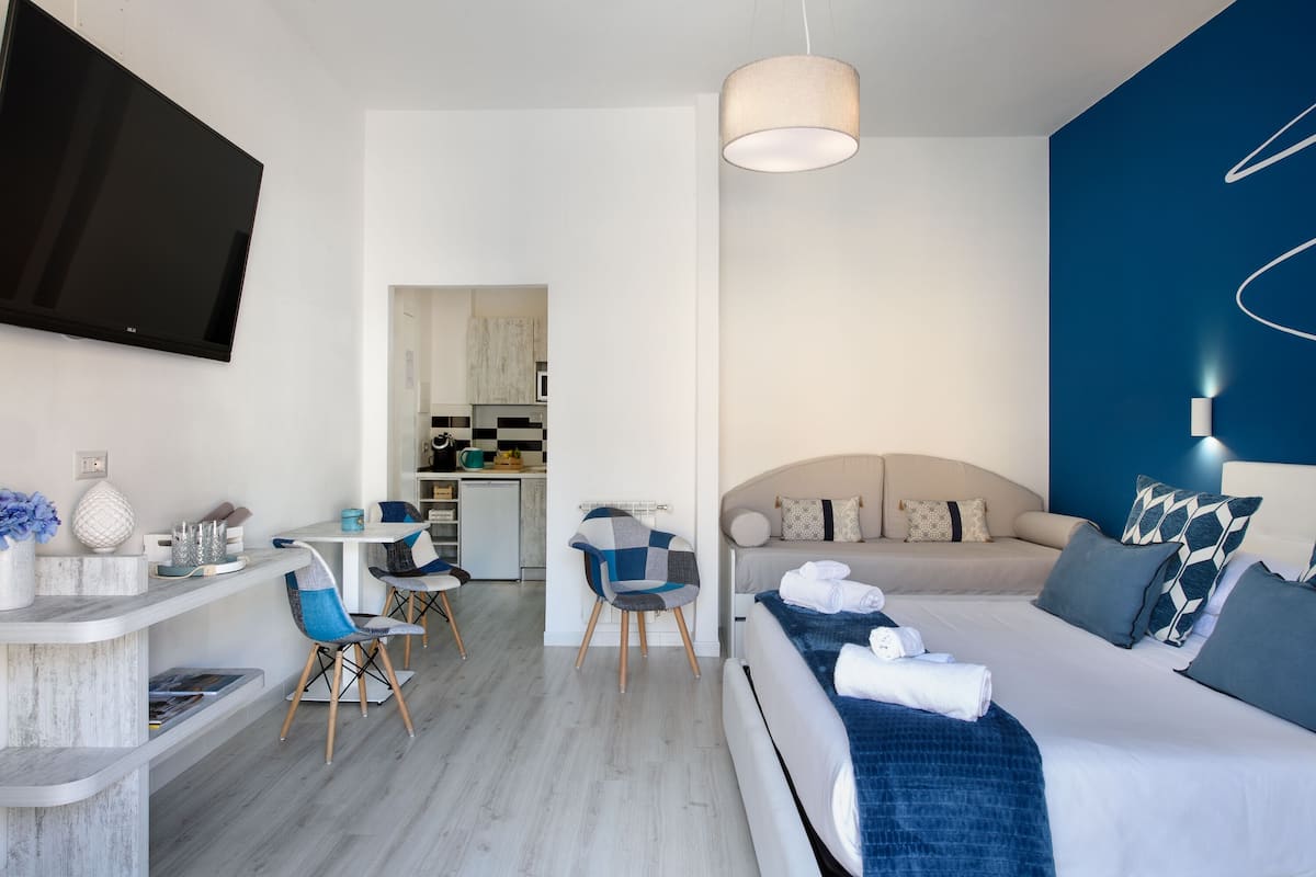 Een moderne hotelkamer met een flatscreen-tv, een bank met blauwe en witte beddengoed, een zithoek met een tafel en stoelen, en een keuken op de achtergrond.