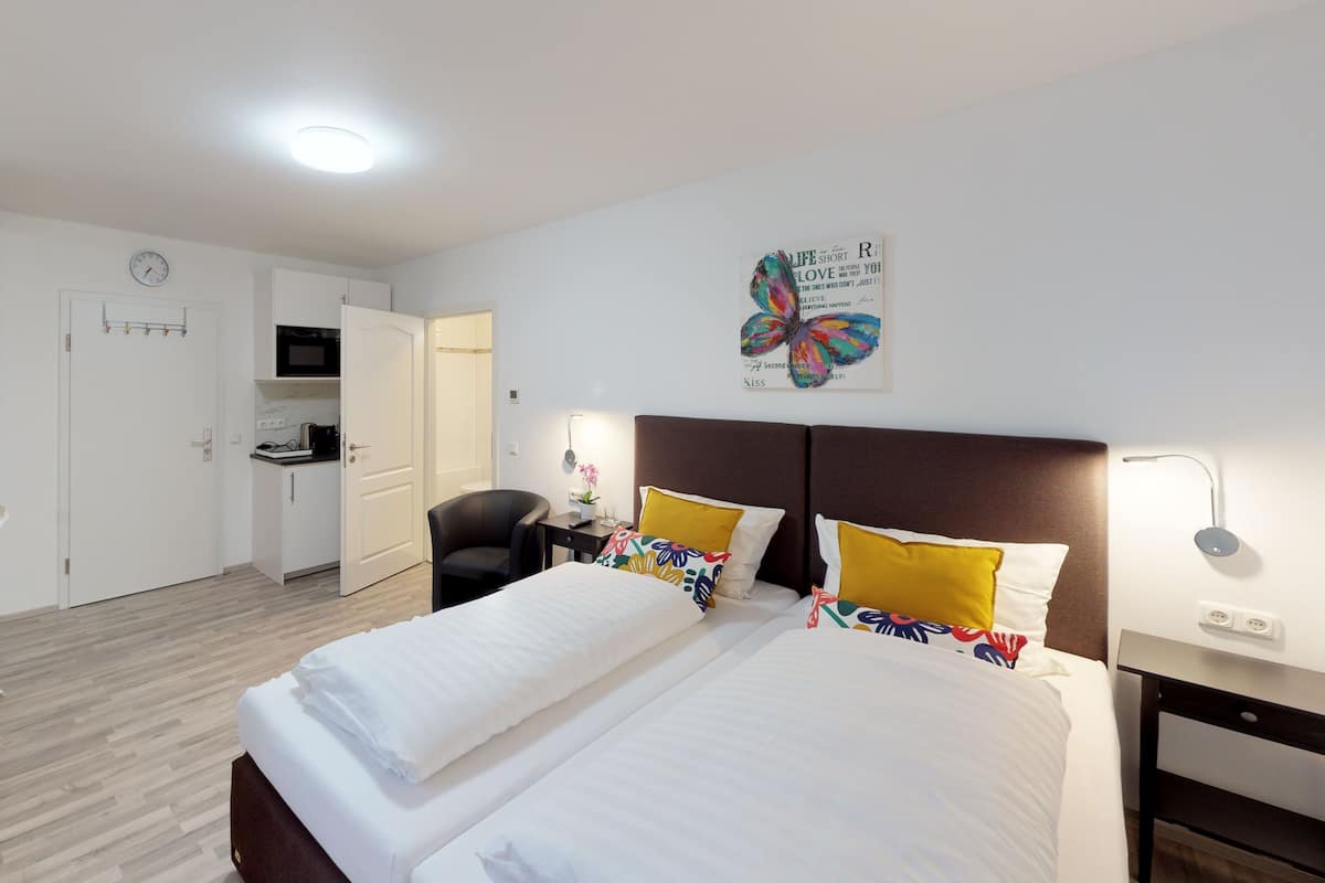 A hotel room with a bed, bedside tables, a desk with a chair, a small kitchenette, and a clock on the wall.