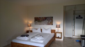 A neatly made bed with white linens and towels, two bedside lamps, a painting on the wall, and a wardrobe with a hanging rack.