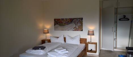 A neatly made bed with white linens and towels, two bedside lamps, a painting on the wall, and a wardrobe with a hanging rack.