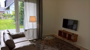 A living room with a sofa, a floor lamp, a glass table, a TV on the wall, and a view of the outdoors.
