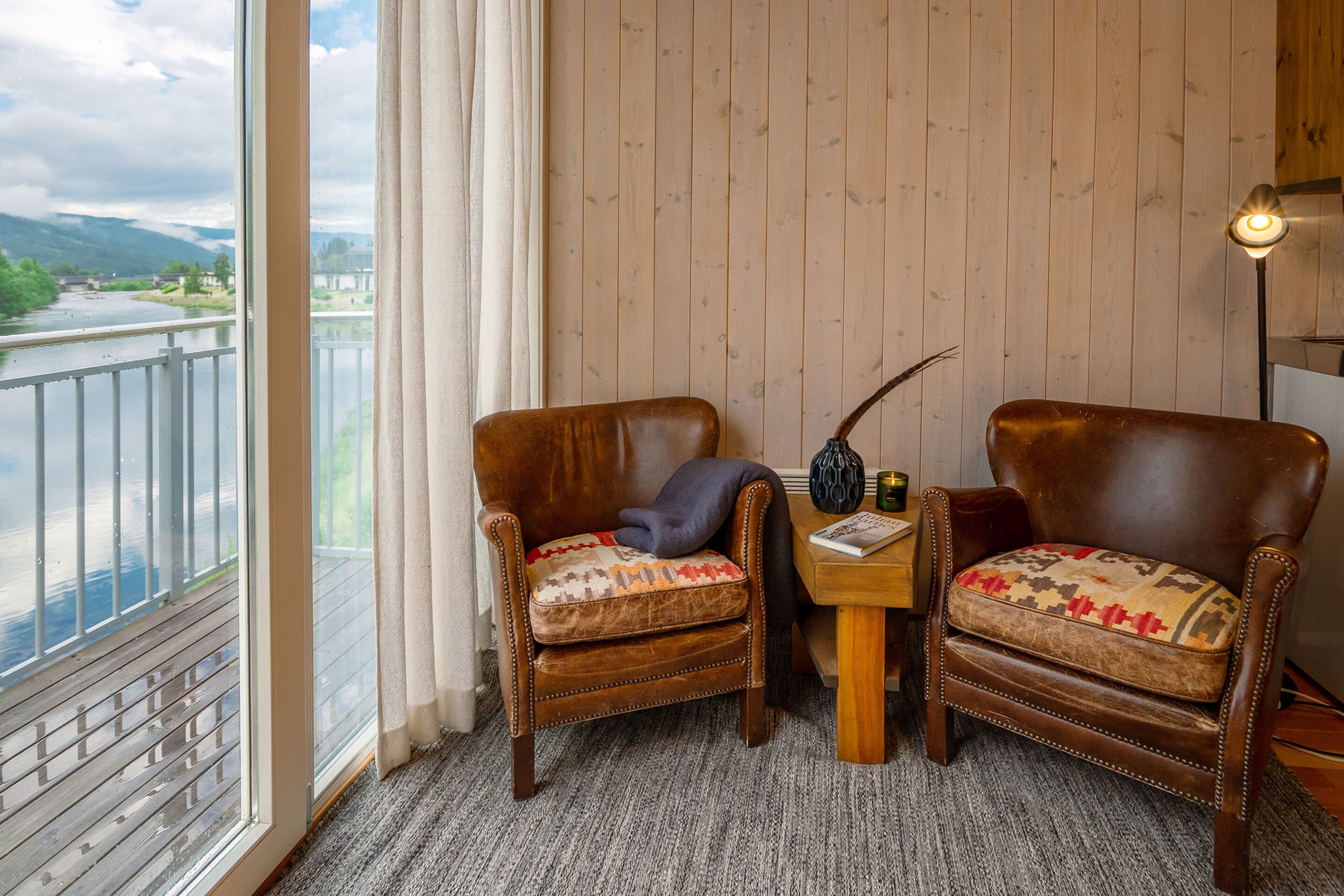 Quarto de canto com duas poltronas de couro, uma pequena mesa de madeira e vista para um rio e montanhas através de uma grande janela.