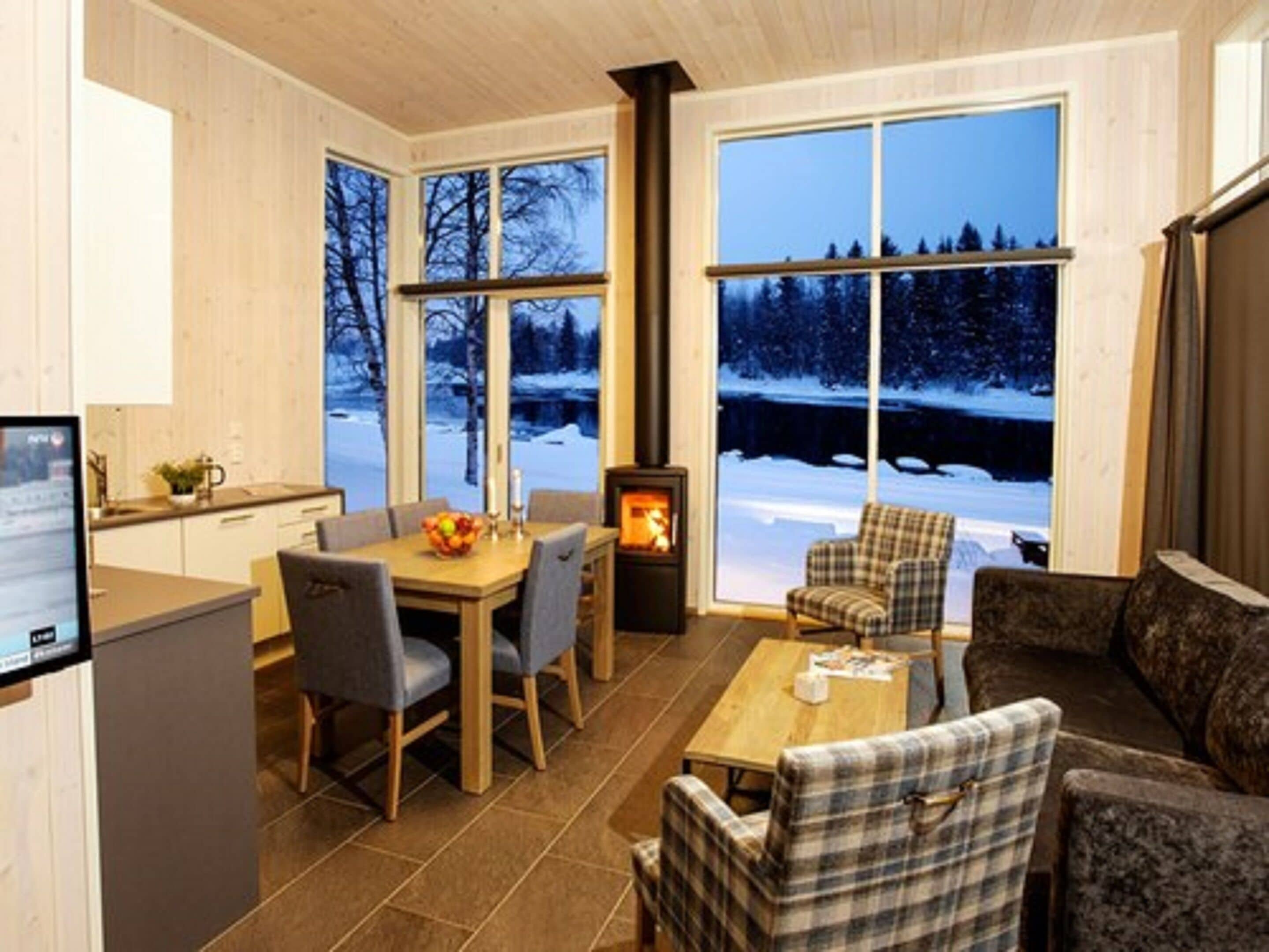 Een knus vertrek met een open haard, een eettafel met stoelen en grote ramen met uitzicht op een besneeid landschap.
