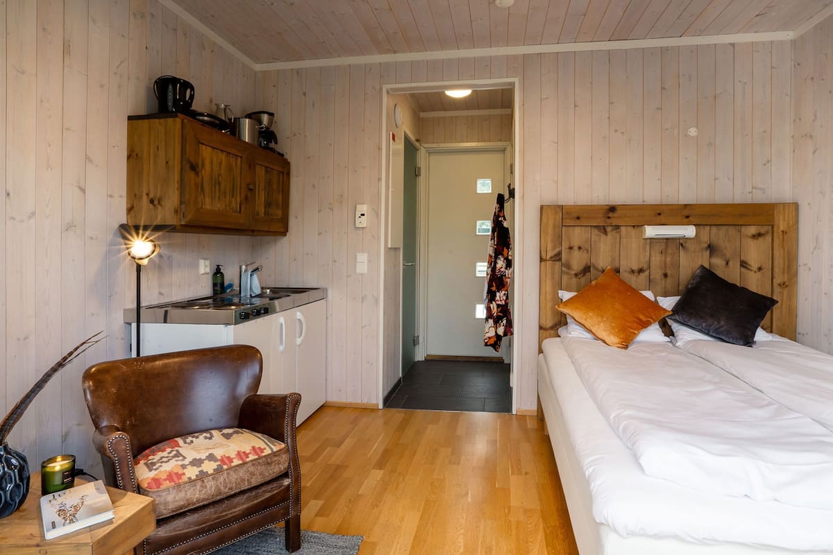 Quarto com cama, cabeceira de madeira, uma cadeira, escrivaninha e cozinha compacta.