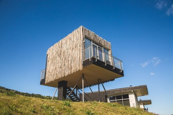 A high-rise wooden structure with a balcony, supported by metal beams, overlooking a grassy area.
