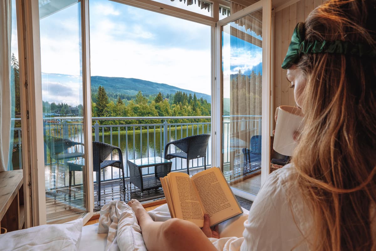 Alguém lendo um livro numa varanda com vista para um lago e montanhas.