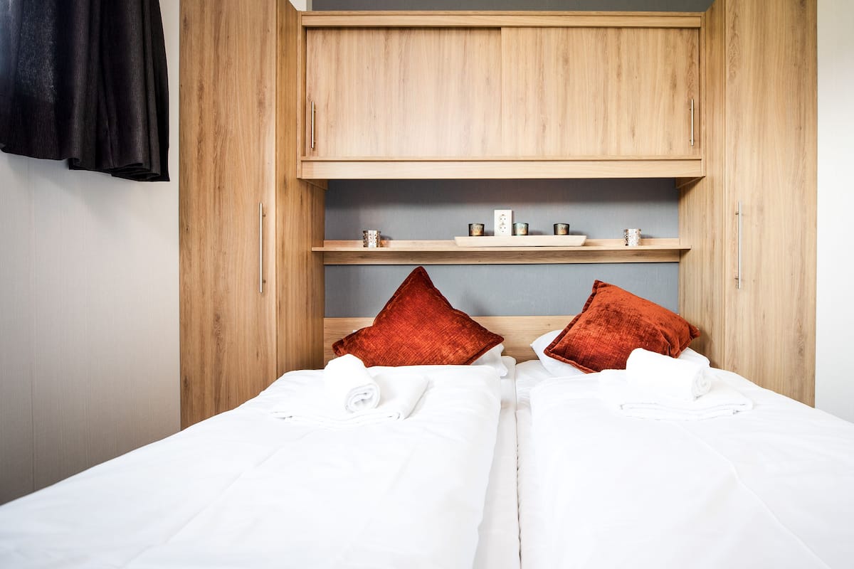 Cama de casal com lençóis brancos e duas almofadas vermelhas, guarda-roupa de madeira com prateleira superior e pequena estante com velas e tomada.