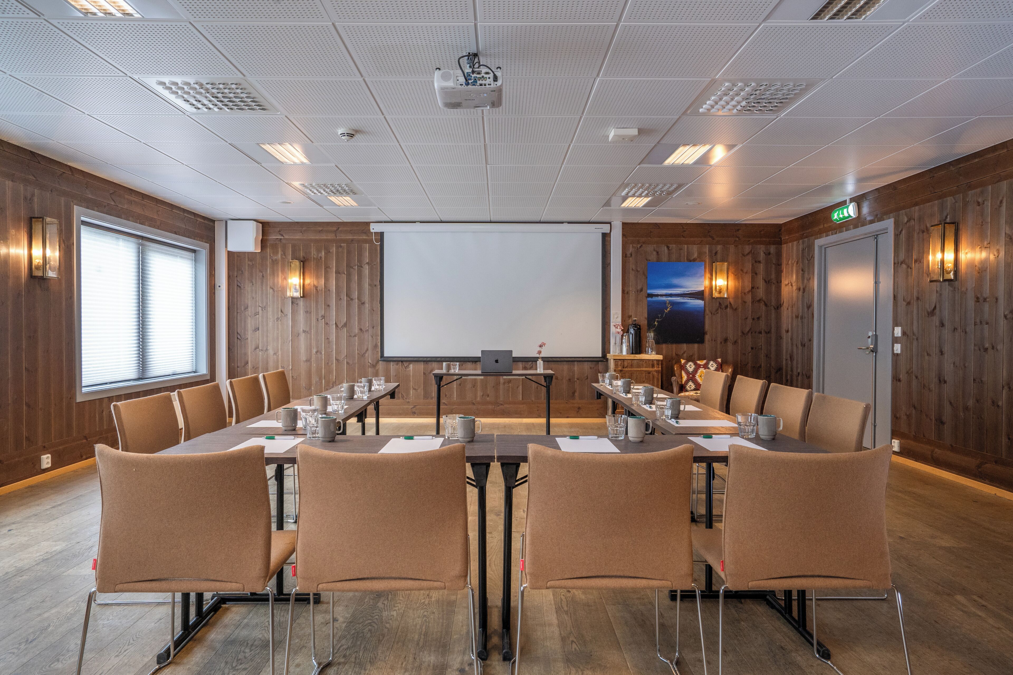 Una sala de conferències amb una taula gran, cadires, un projector, una pantalla i una paret de fusta.