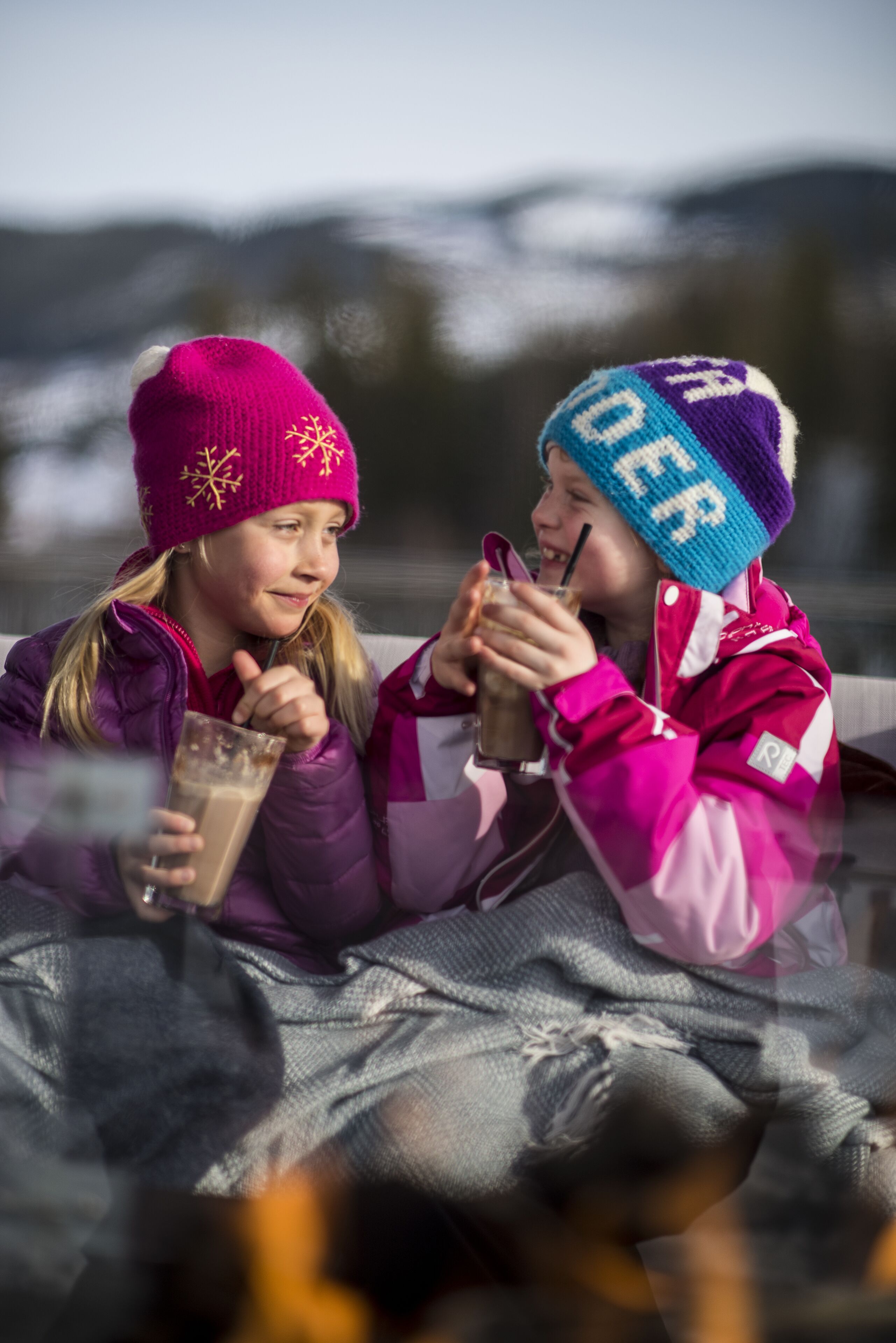 Dos personas sentadas en un trineo, con gorros y abrigos de invierno, sosteniendo bebidas.
