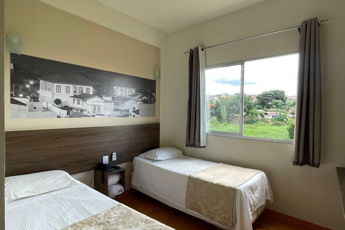 A hotel room with two beds, a nightstand, a large window with curtains, and a wall-mounted painting of a building.