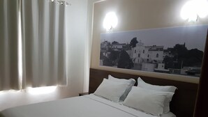 Una habitación de hotel con una cama, cortinas blancas y una gran foto enmarcada de un paisaje urbano en la pared.