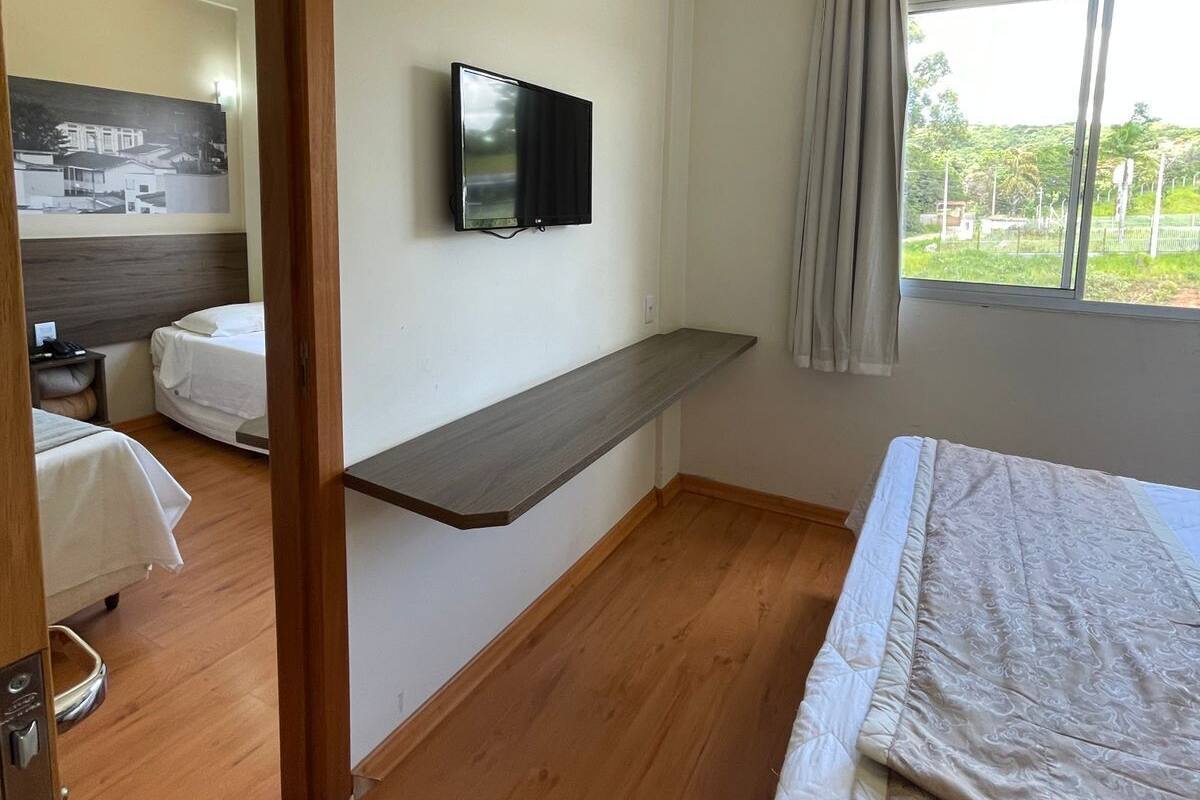 A hotel room with a bed, a TV mounted on the wall, a window with curtains, and a wooden floor.