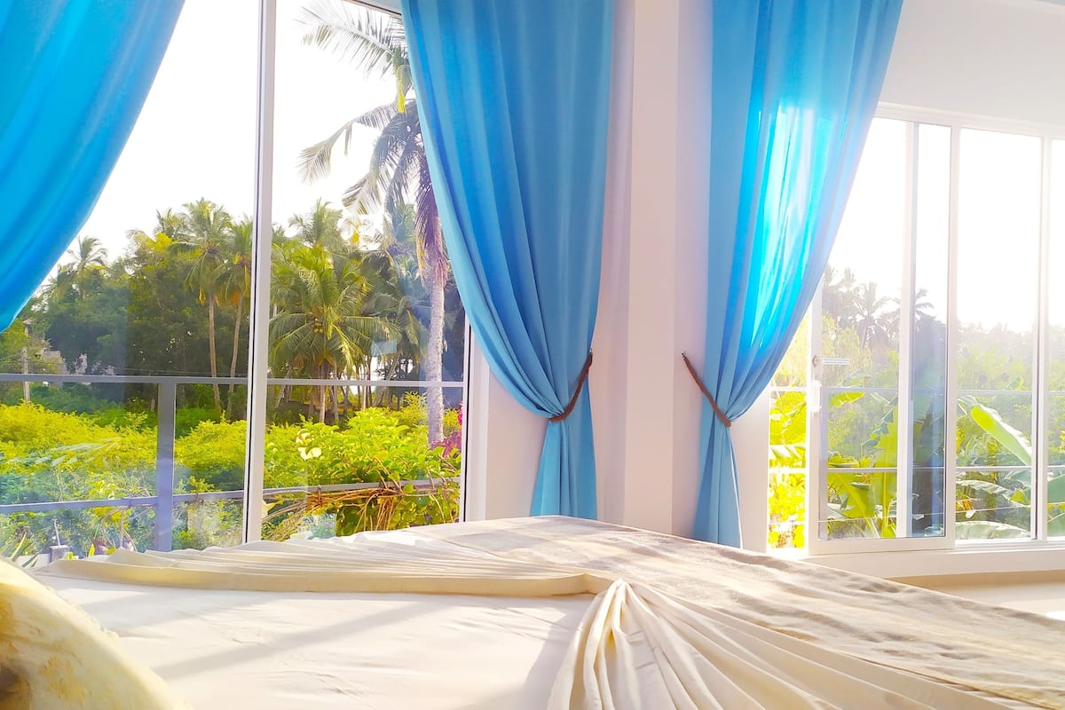 A room with a large window, curtains, a bed, and a view of greenery outside.