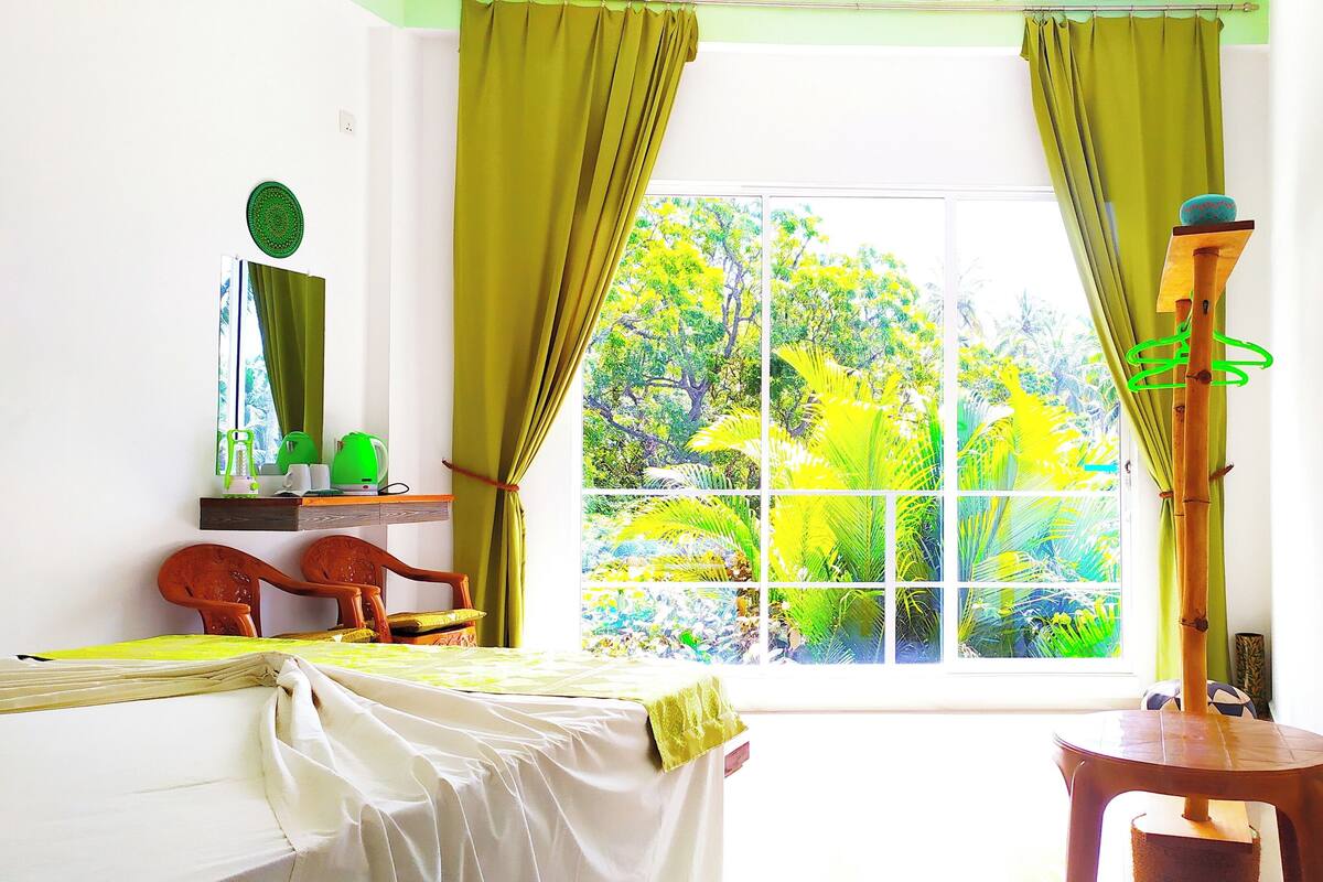 A room with a bed, a chair, a table, a window with curtains, and a view of greenery outside.
