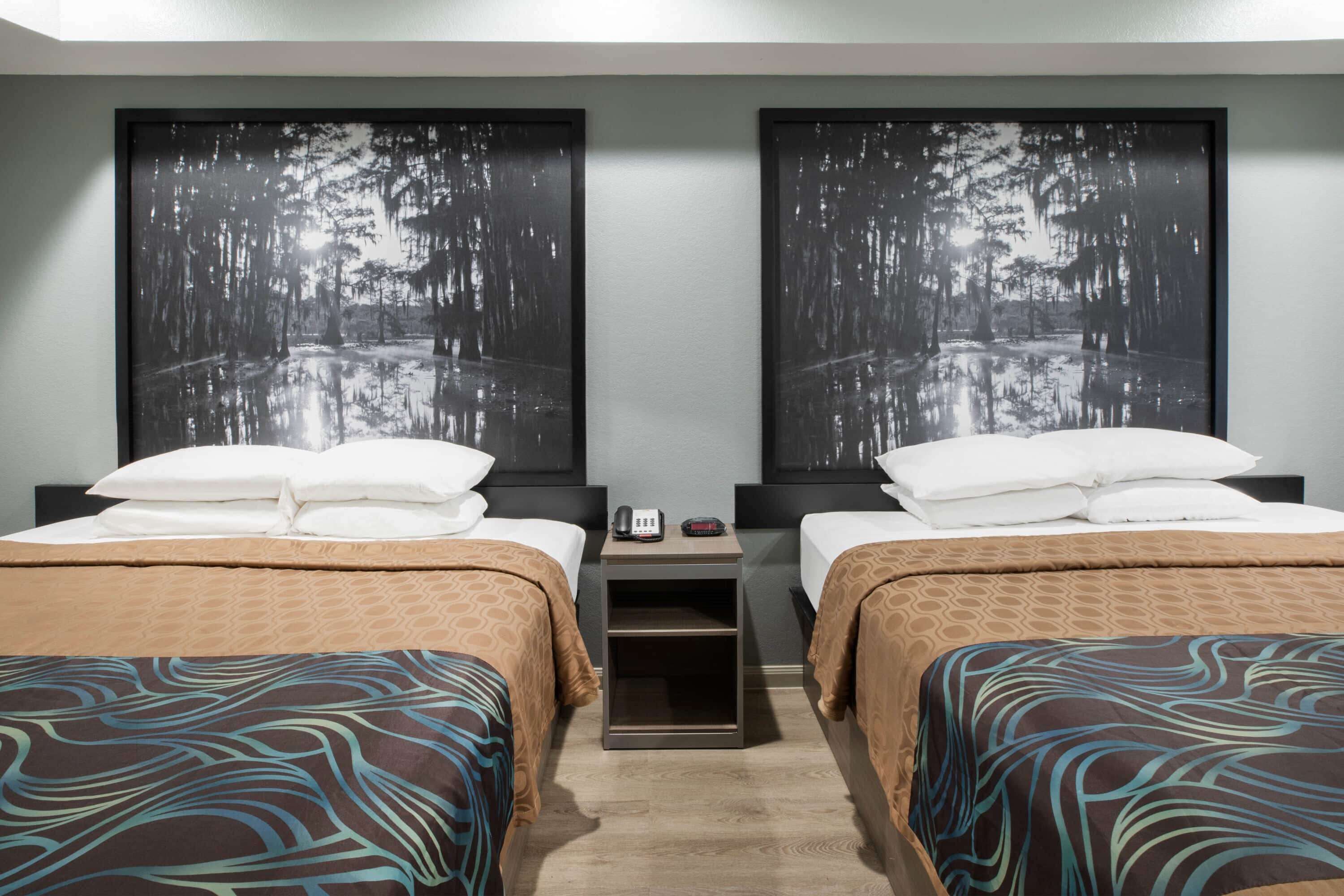A hotel room with two beds, each with a patterned bedspread, a nightstand with a phone and a clock, and two large framed pictures of a forest scene on the wall.