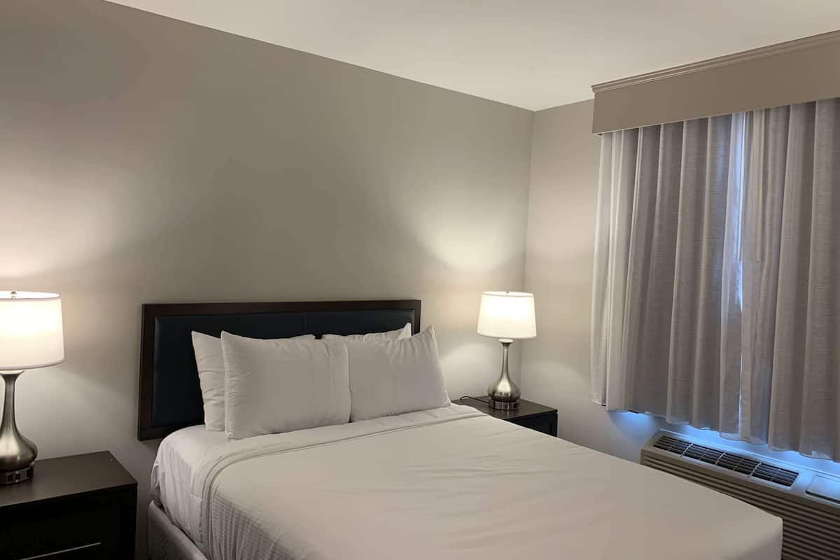 Ein Hotelzimmer mit einem Bett, zwei Nachttischlampen, einem Fenster mit Vorhängen und einem Wandlüfter.