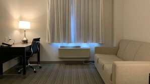 A hotel room with a desk, chair, lamp, air conditioner, and a sofa. - Wingate by Wyndham JFK Airport/Far Rockaway (Far Rockaway)