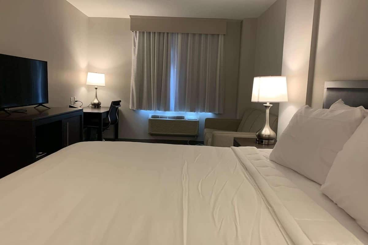 Ein Hotelzimmer mit einem großen Bett, einem Schreibtisch mit Flachbildfernseher, einem Sessel, einer Lampe und einem Fenster mit Vorhängen.