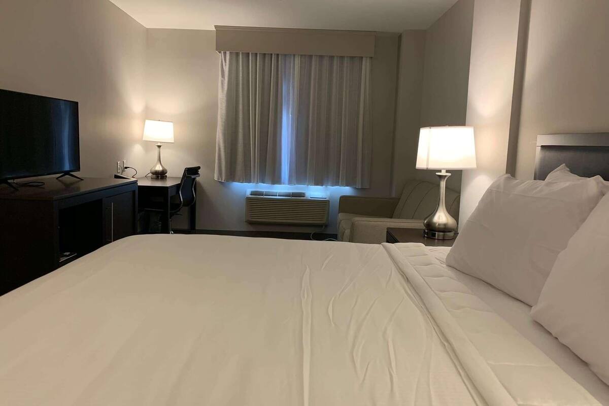 Ein Hotelzimmer mit einem großen Bett, einem Schreibtisch mit Flachbildfernseher, einem Sessel, einer Lampe und einem Fenster mit Vorhängen.