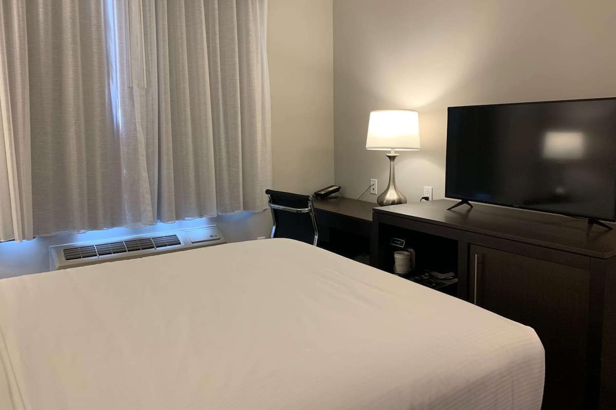 Ein Hotelzimmer mit einem großen Bett, einem Schreibtisch mit Lampe, einem Fernseher und einem Fenster mit Vorhängen.