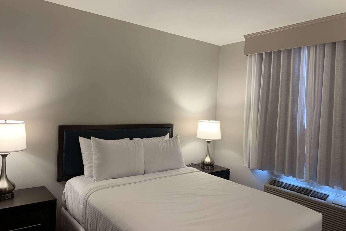 Ein Hotelzimmer mit einem Bett, zwei Nachttischlampen, einem fensterbelaubten Fenster und einer Wand-Klimaanlage.