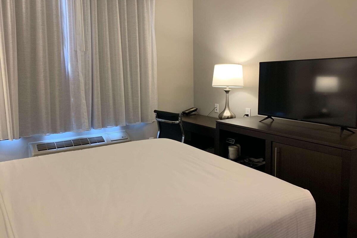 Une chambre d’hôtel avec un grand lit, un bureau avec une lampe, une télévision et une fenêtre avec des rideaux.