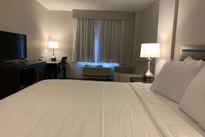 A hotel room with a large bed, a desk with a flat-screen TV, a chair, a lamp, and a window with curtains. - Wingate By Wyndham Jfk Airport Far Rockaway (Far Rockaway)
