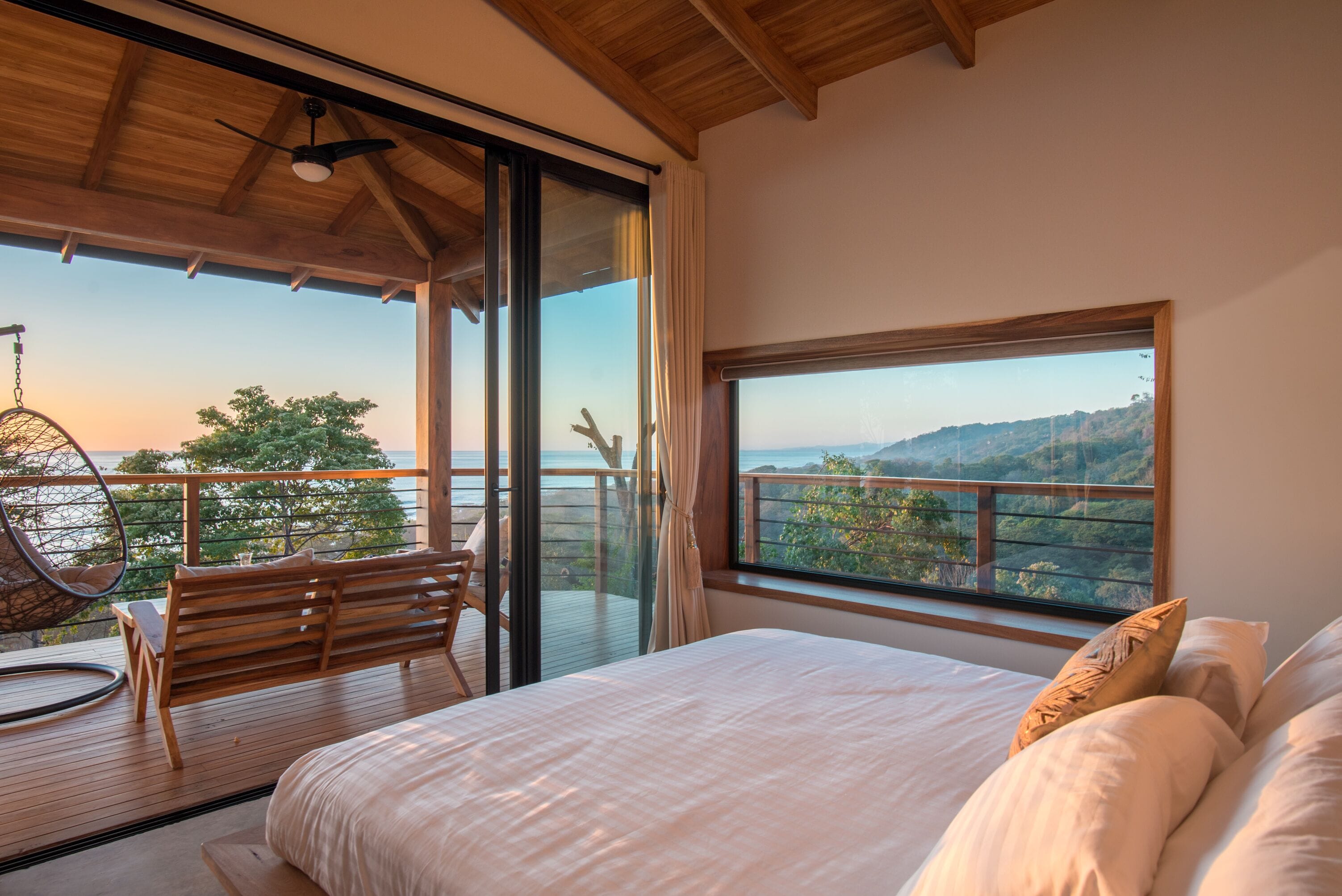 Un dormitorio con una cama grande, un banco de madera y una silla colgante en un balcón con vistas panorámicas.