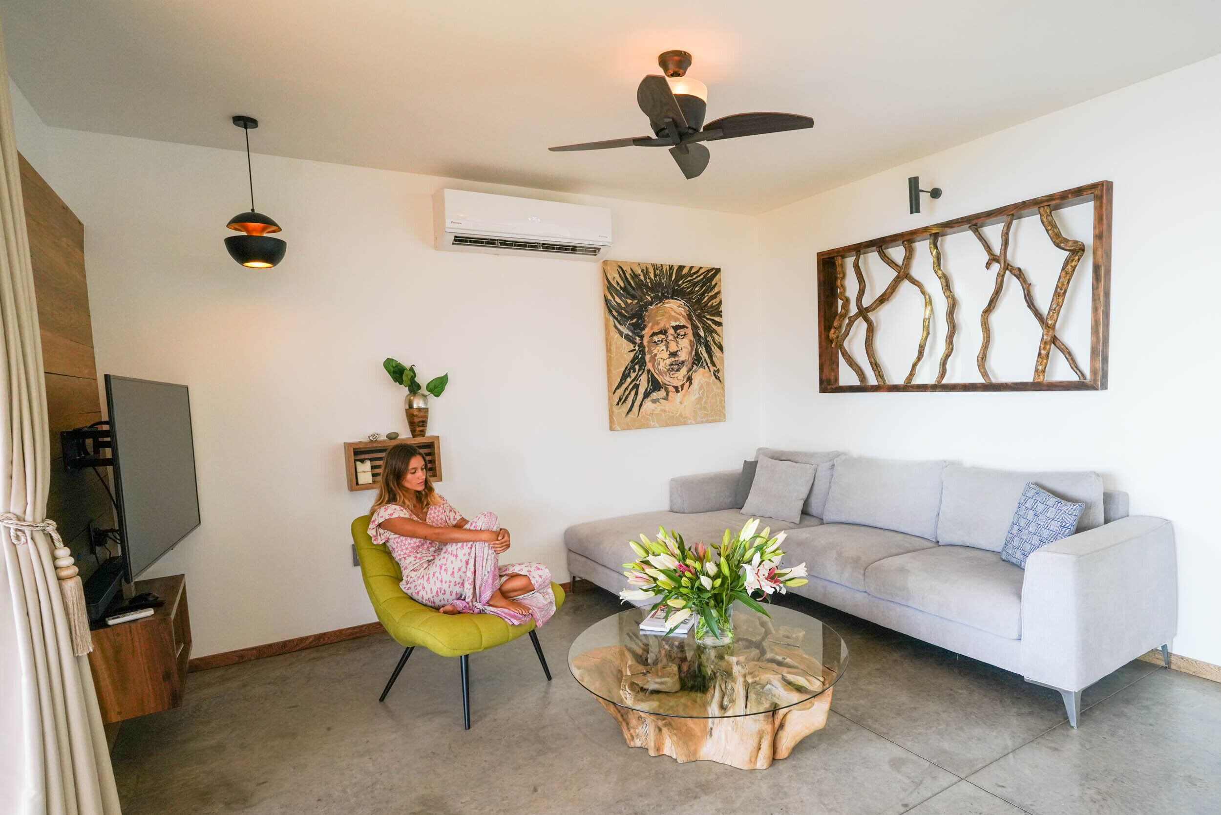 Una sala de estar con un sofá gris, una mesa de centro de vidrio con flores, un ventilador de techo y un cuadro en la pared.