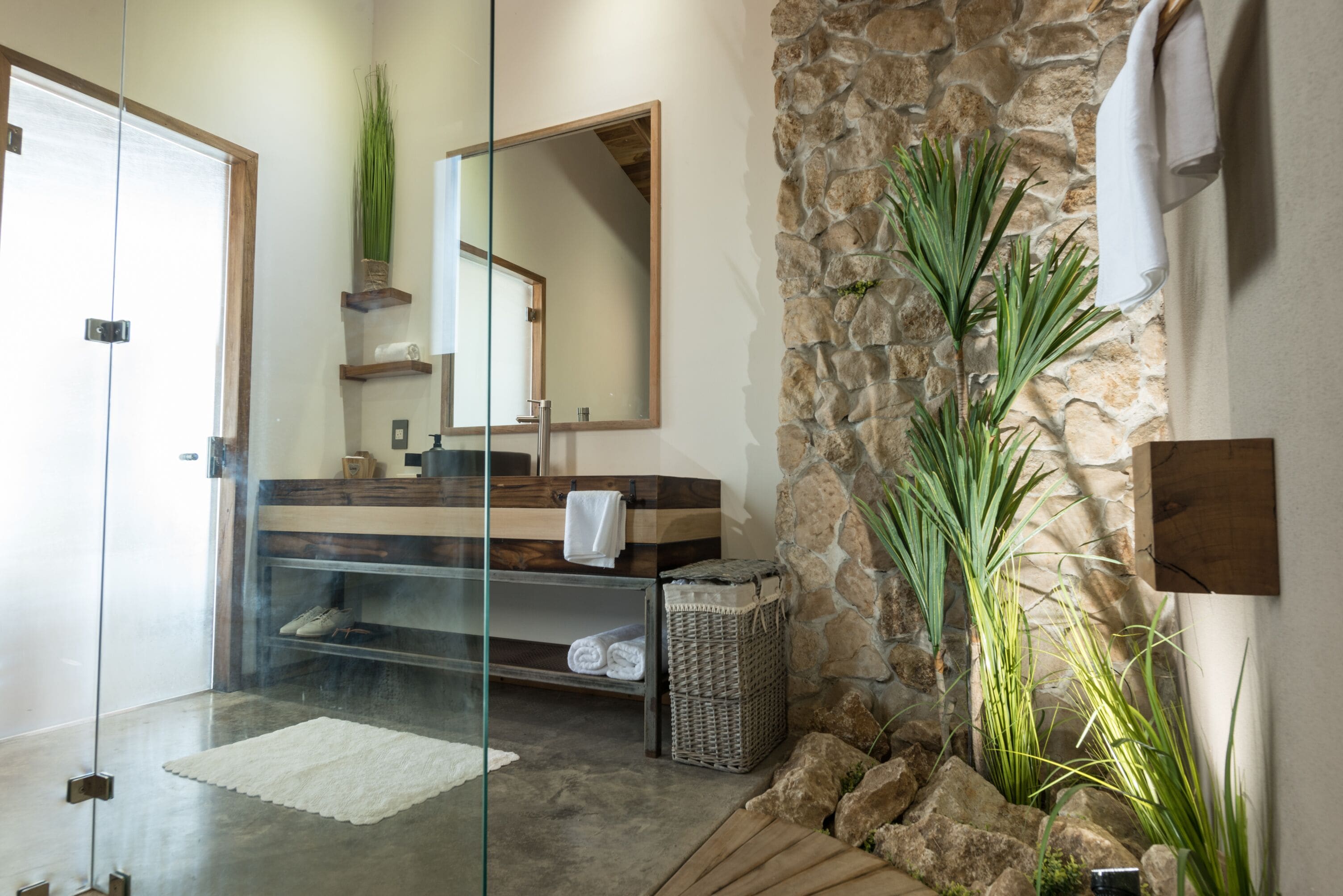 A modern bathroom with a glass shower, stone wall, and a wooden vanity.