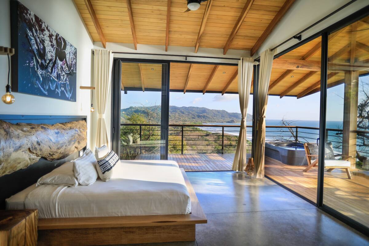 Un dormitorio moderno con una cama grande, cabecera de piedra y un cuadro en la pared. Hay un balcón con vistas al océano y las colinas.