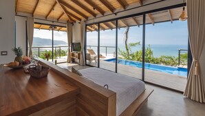 Een moderne slaapkamer met een groot bed, houten plafond en uitzicht op een overloopzwembad en de oceaan.