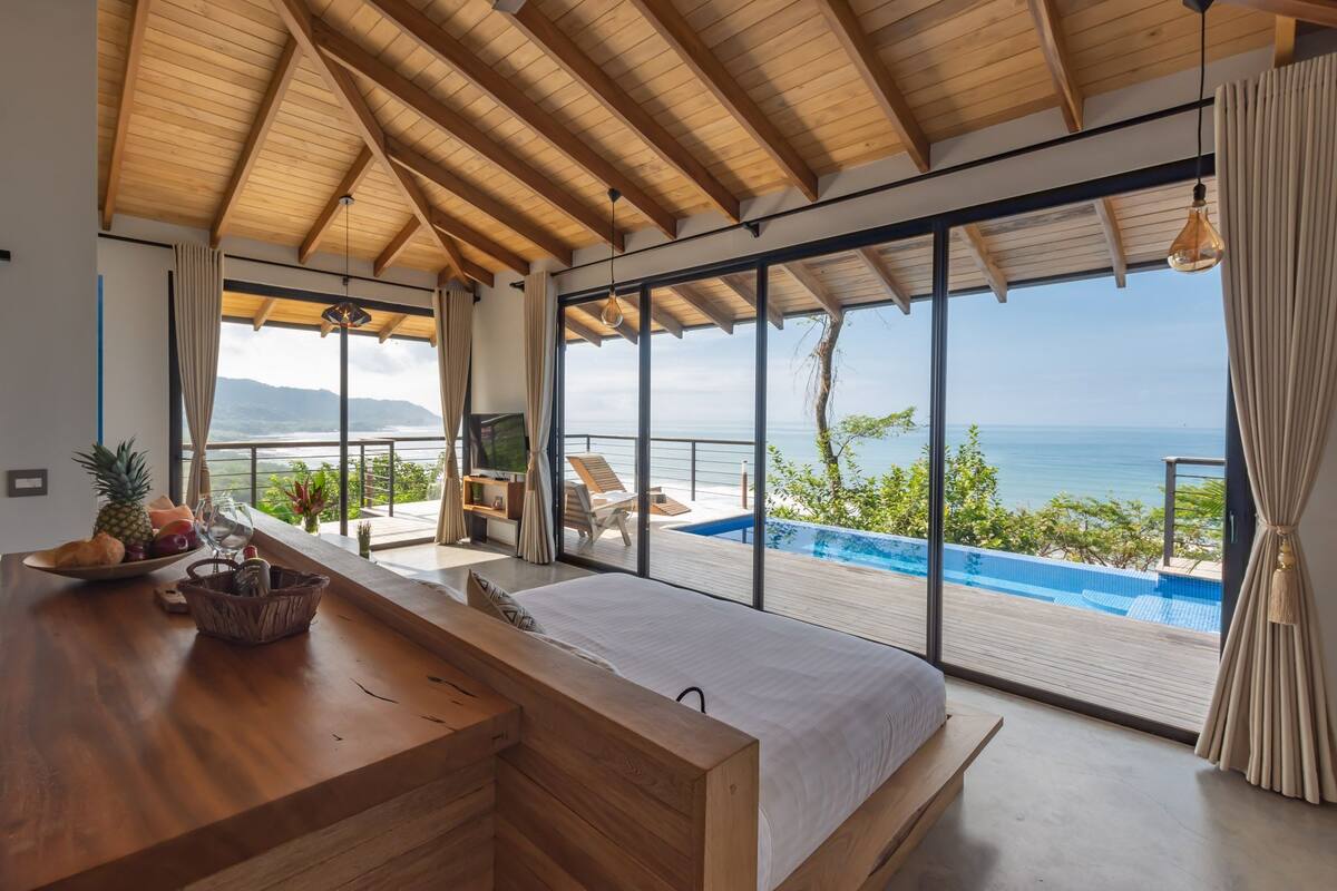 Un dormitorio moderno con una cama grande, techo de madera y vistas a una piscina infinita y al océano.