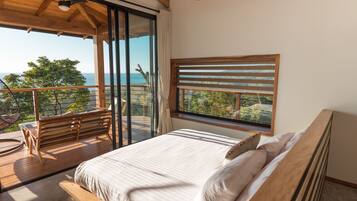 A bedroom with a large bed, a wooden bench, and a view of the sea through a window.